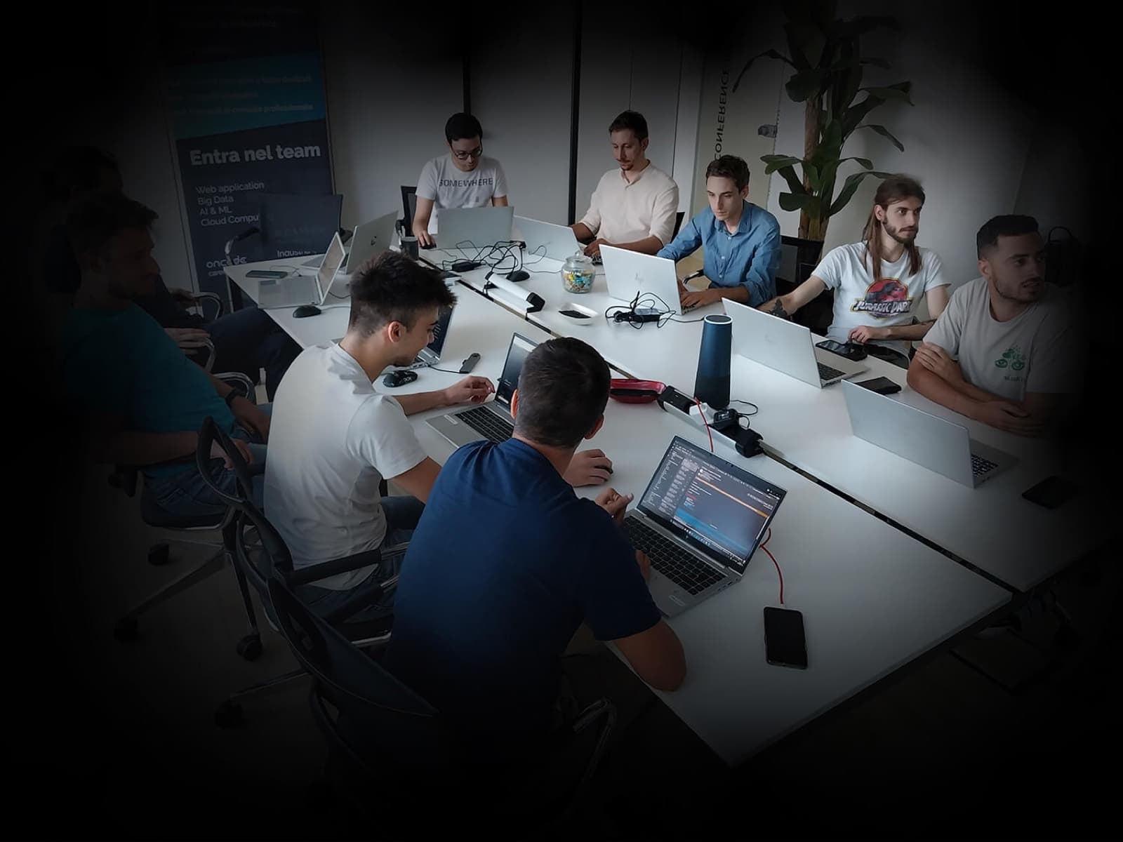Otto colleghi attorno a un tavolo lungo con i portatili aperti, in sala riunioni, accanto a un roll-up «Entra nel team»