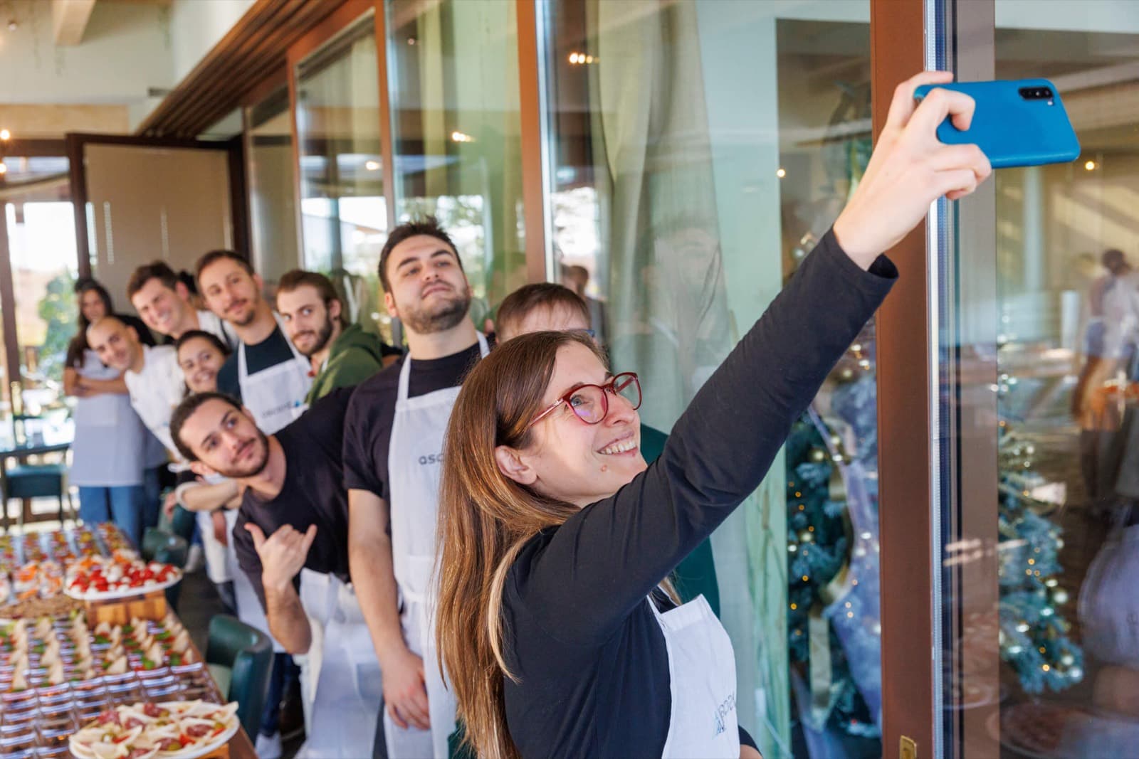 Colleghi in grembiule a un evento di cucina aziendale, una di loro scatta un selfie davanti al buffet